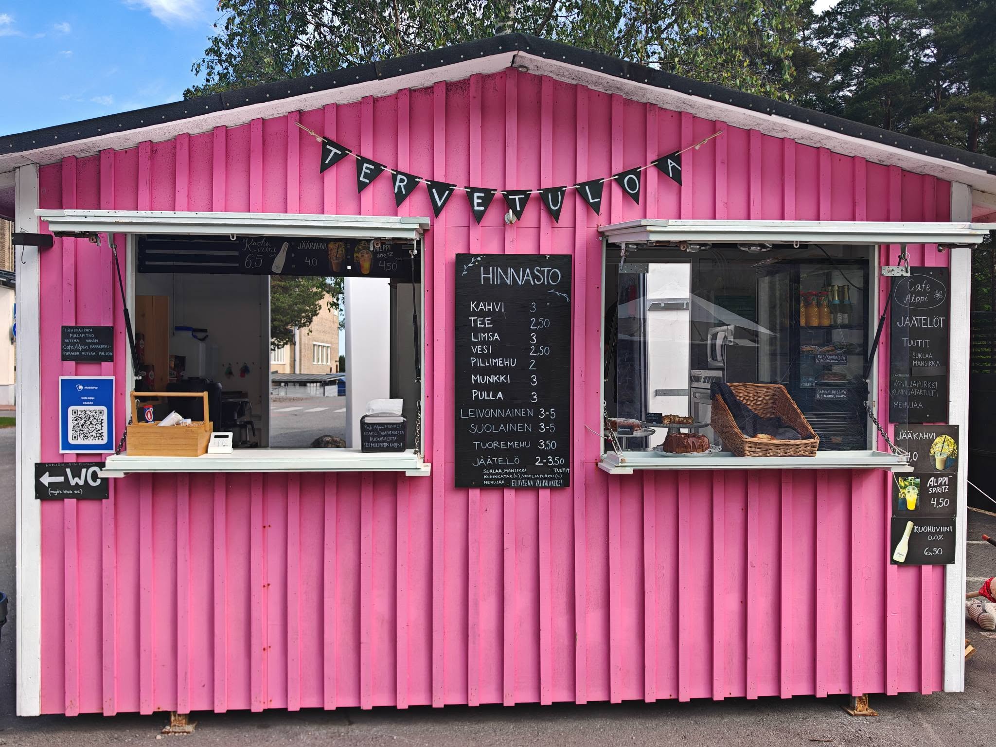 Cafe Alpin pinkki kioski. Kaksi avattua myyntiluukkua, niiden välissä käsin kirjoitettu hinnastotaulu, ja katonrajassa TERVETULOA-viirinauha.