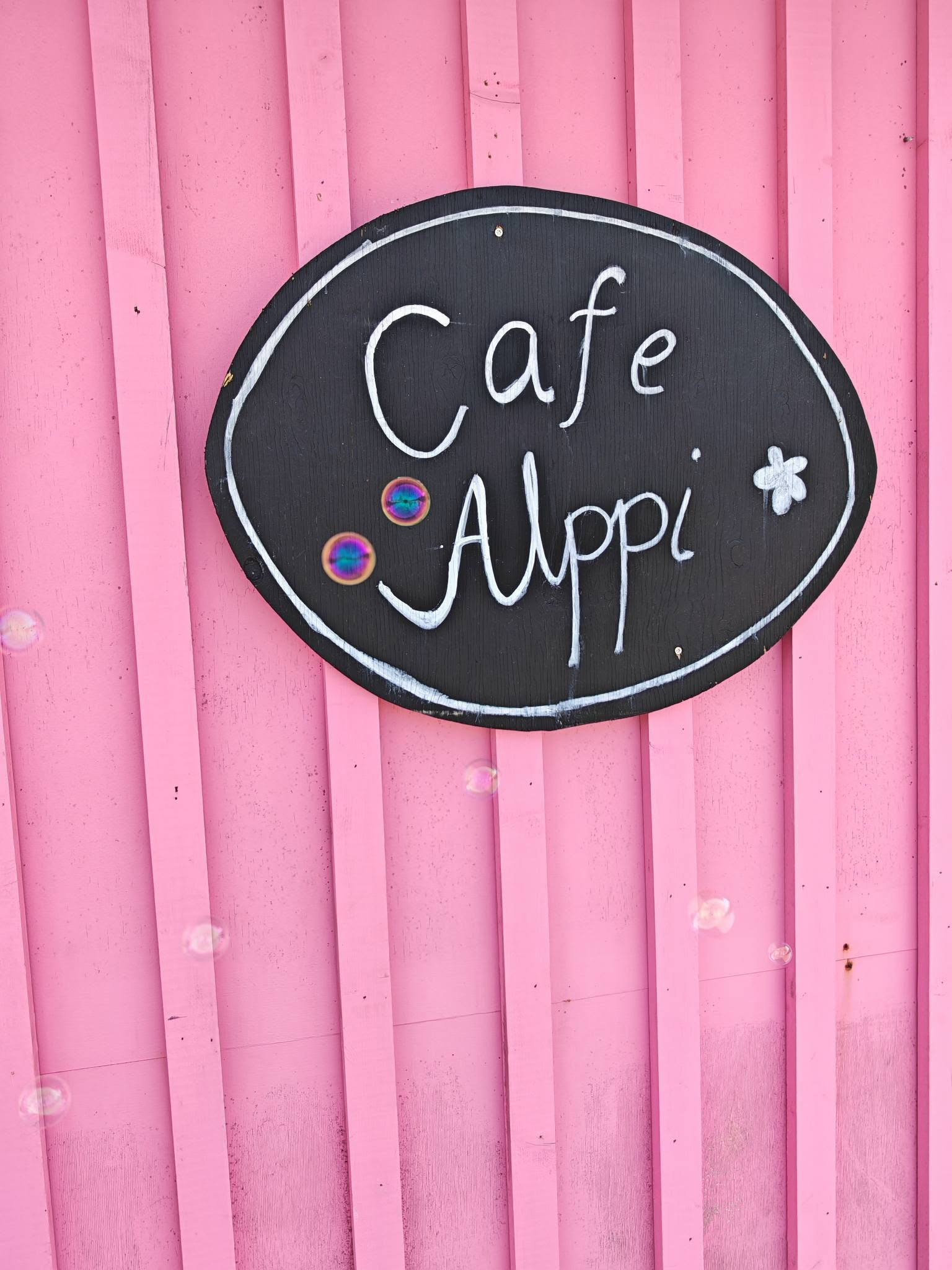 Musta soikea kyltti, johon on maalattu valkoisella käsialalla Cafe Alppi. Kyltti roikkuu pinkillä profiilipeltiseinällä, ilmassa saippuakuplia.